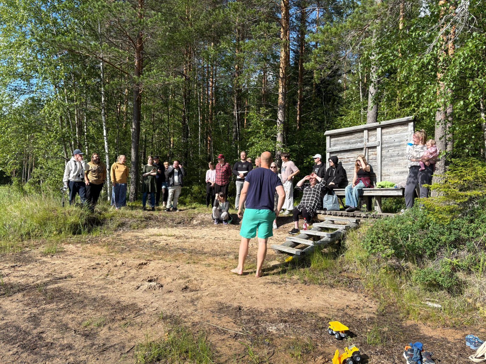 Kastetilaisuus rannalla: Joonas seisoo veden äärellä selkä kameraan, ihmiset kokoontuneet rantaan